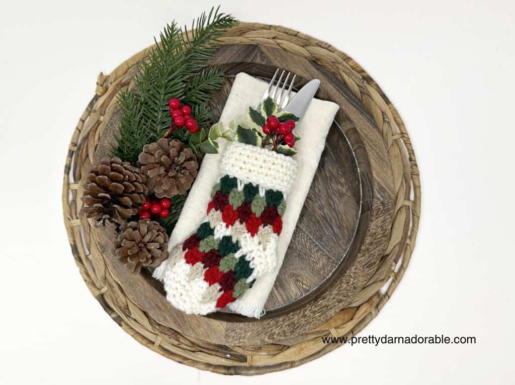 Festive holiday table setting with pinecones, greenery, red berries, and utensils in a crocheted red, green, and white holder; website text in corner. Free crochet pattern