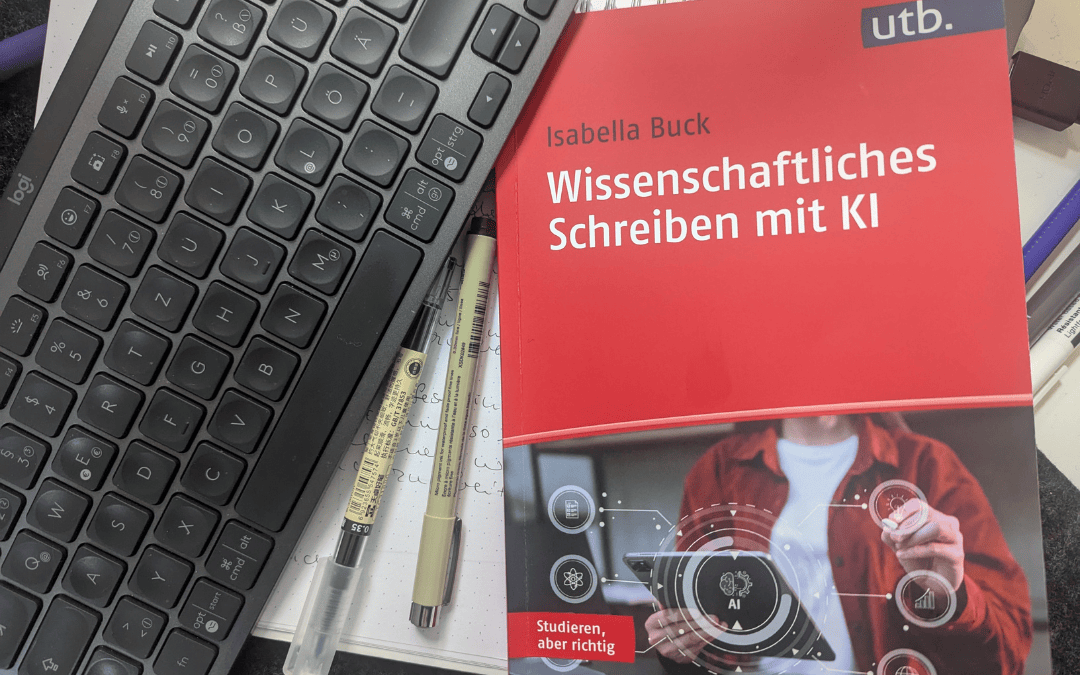 Buch ‚Wissenschaftliches Schreiben mit KI‘ von Isabella Buck liegt auf einem Schreibtisch neben einer Tastatur, zwei Stiften und einem Notizbuch mit handschriftlichen Notizen.