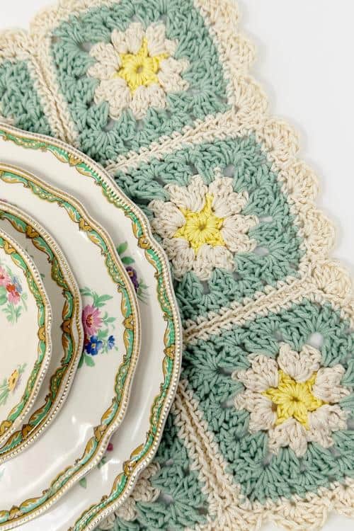 Elegant floral-patterned plates are stacked on a crochet placement with green, cream, and yellow square patterns, perfect for adding charm to your kitchen decor against a white background.