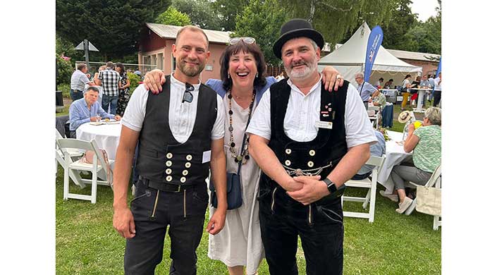 Drei lachende Erwachsene stehen draußen zusammen, zwei Männer in traditioneller Kleidung und eine Frau in einem weißen Kleid.
