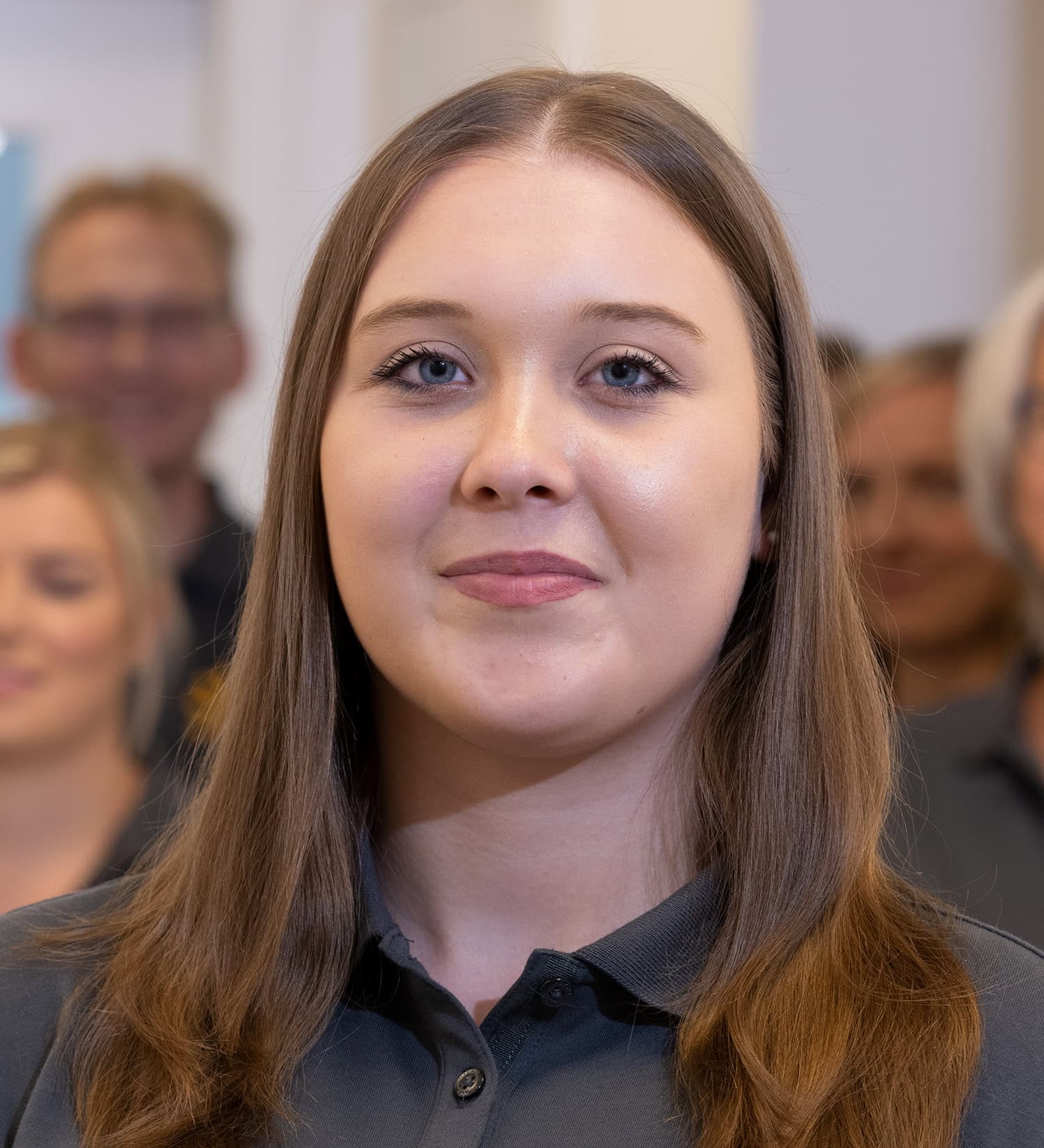Portraitbild einer braunhaarigen Mitarbeiterin in Arbeitskleidung mit dem Praxis-Team im Hintergrund.