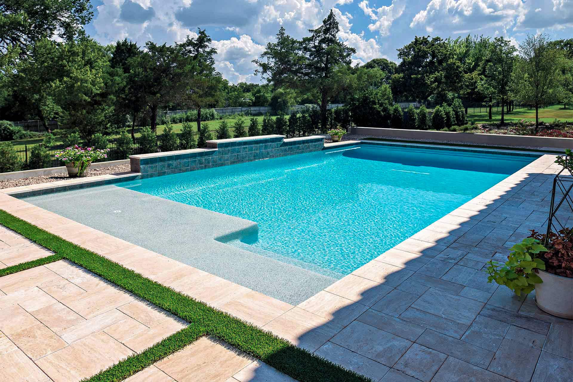 Rectangular outdoor swimming pool with clear blue water, surrounded by stone tiles and lush landscaping. Discover the lasting beauty of New Gunite Pools, set amid trees in the background under a partly cloudy sky.