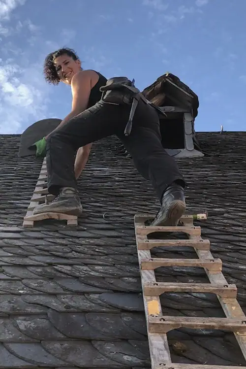 Sonja Theisen arbeitet auf einem Schieferdach und hält eine Schieferplatte, während sie auf einer Holzleiter steht. Der Himmel ist blau mit wenigen Wolken.