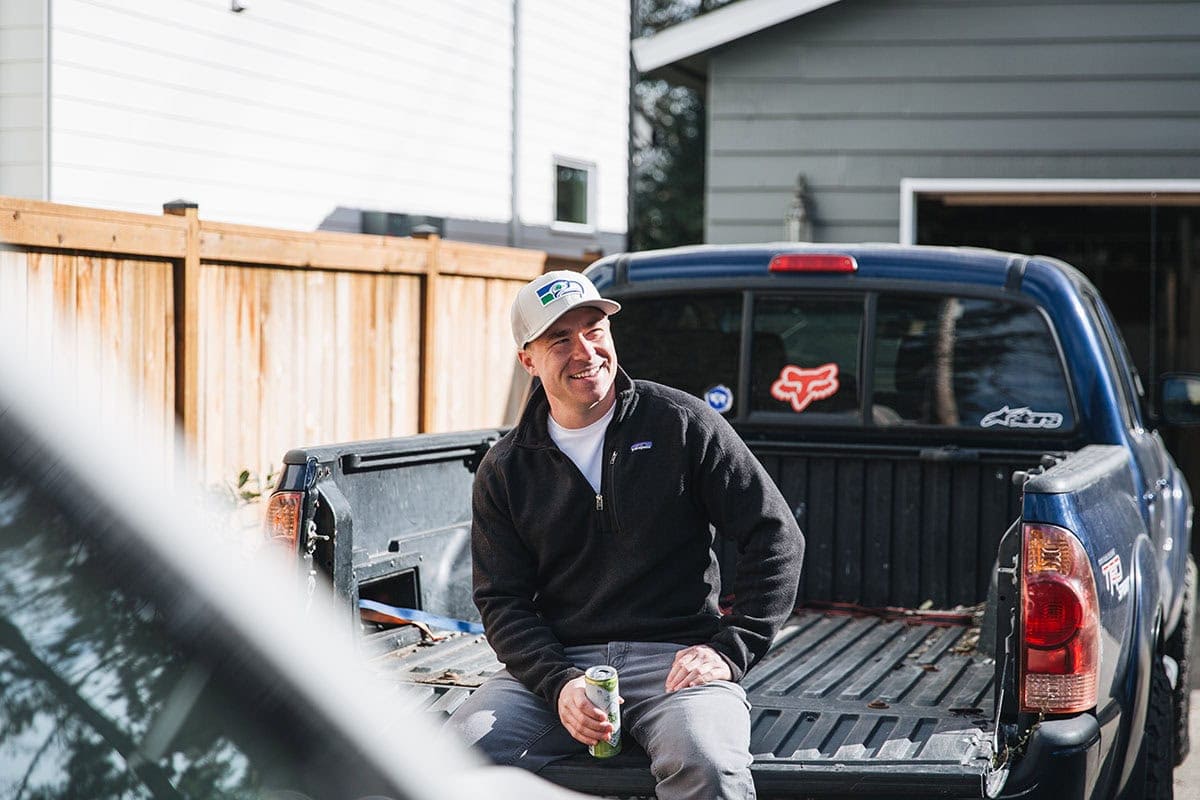 A man in a black jacket and cap sits on the tailgate of a blue pickup truck, holding a can, in a sunny residential backyard.