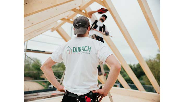 Zwei Bauarbeiter arbeiten an einer Holzrahmenkonstruktion, einer trägt ein DURACH-T-Shirt und eine Mütze, der andere steht auf dem Gerüst im Hintergrund.