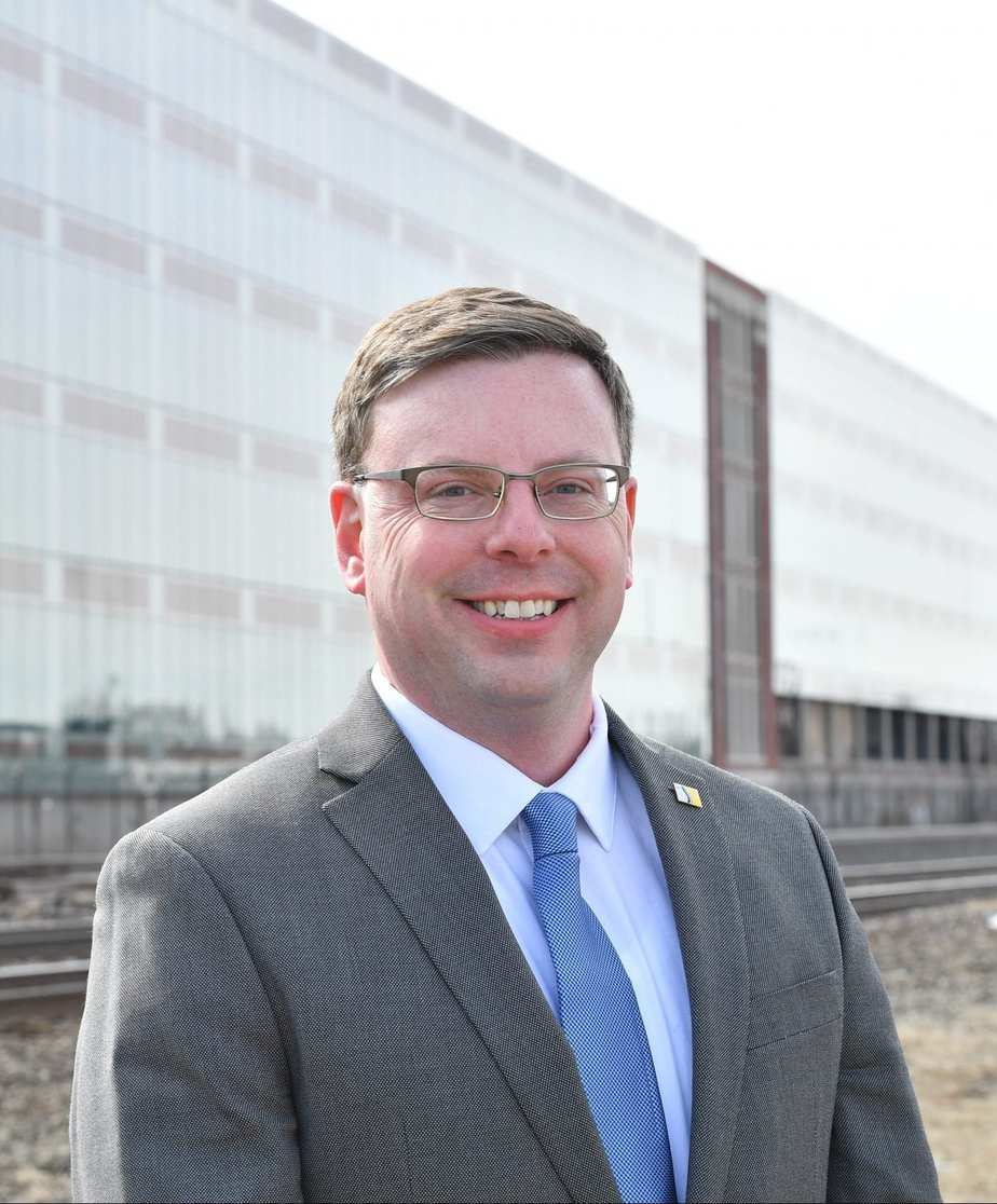 mayor_mueller_bio-scaled-e1578351042118 A professional man in a suit standing outdoors near a modern building with train tracks in the background.