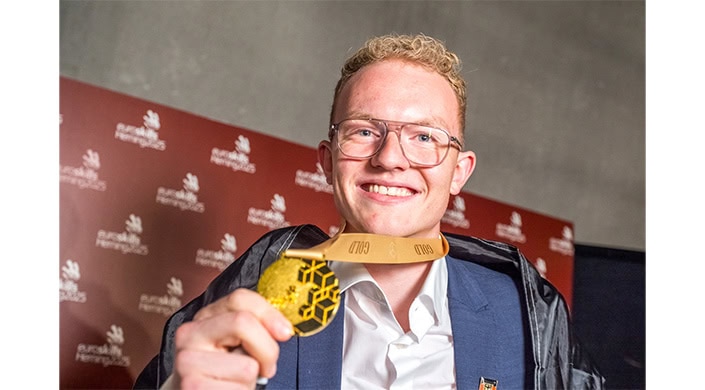 Ein junger Mann mit Brille lächelt und hält eine goldene Medaille.
