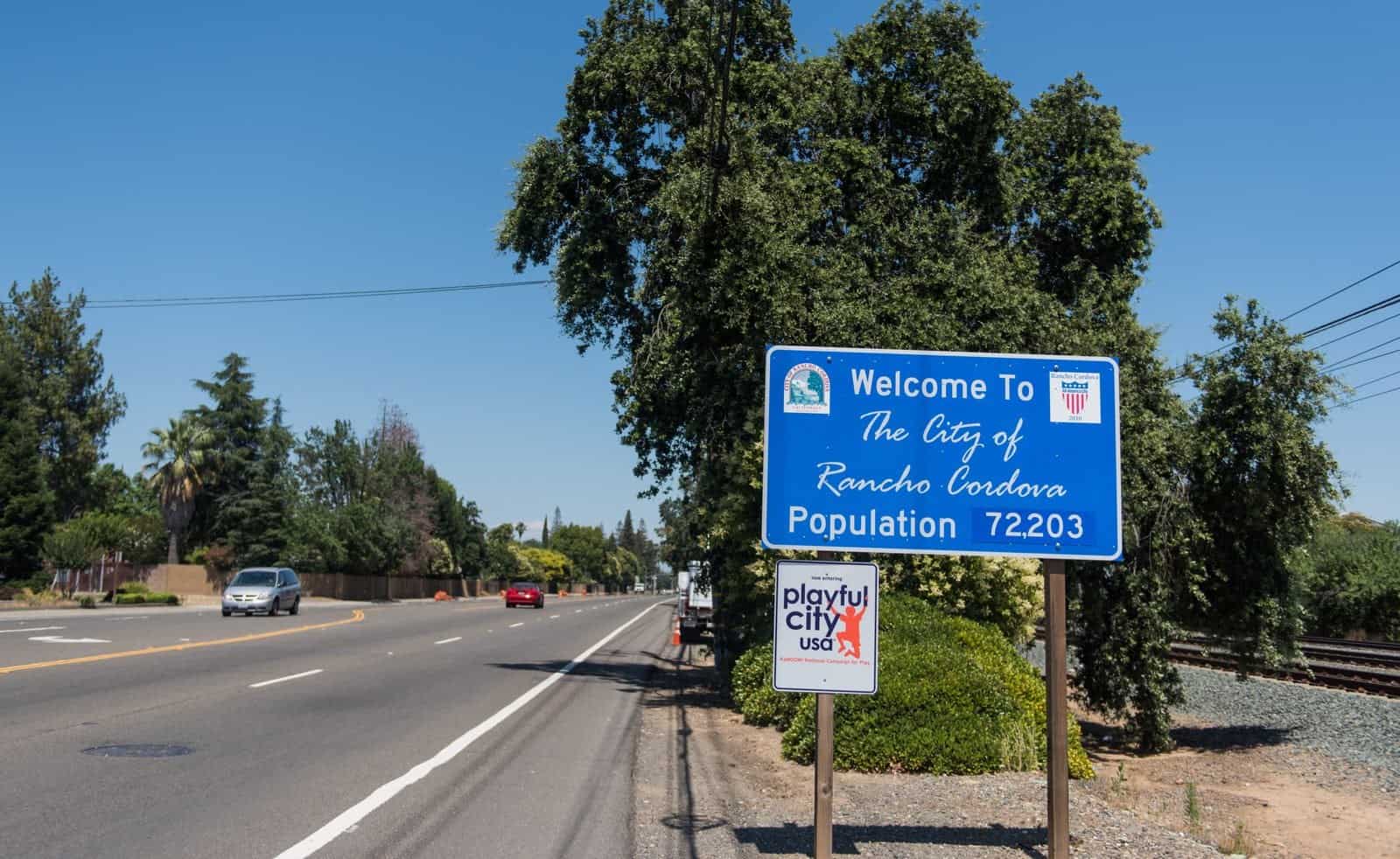 A blue Welcome to the City of Rancho Cordova sign beside a suburban road lined with trees and power lines.