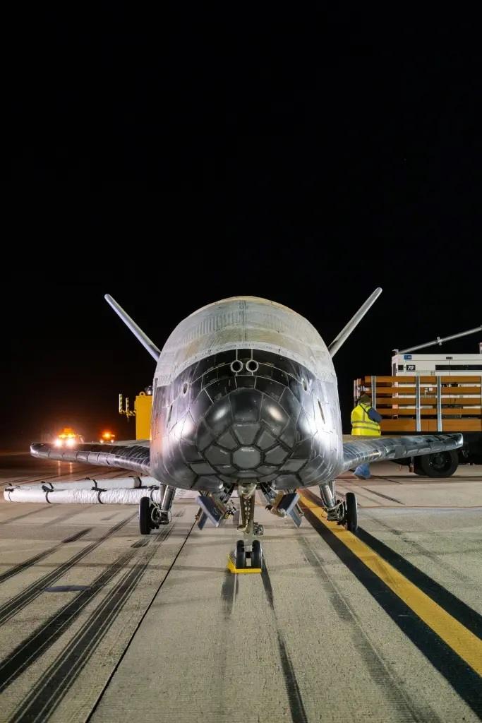 The X-37B Space Force orbital vehicle, a miniature space shuttle, rests on the Vandenberg landing strip at night, illuminated by ground lights. The unmanned spacecraft, part of a secretive Space Force mission, is surrounded by ground crew and equipment, showing its return after a long space mission.