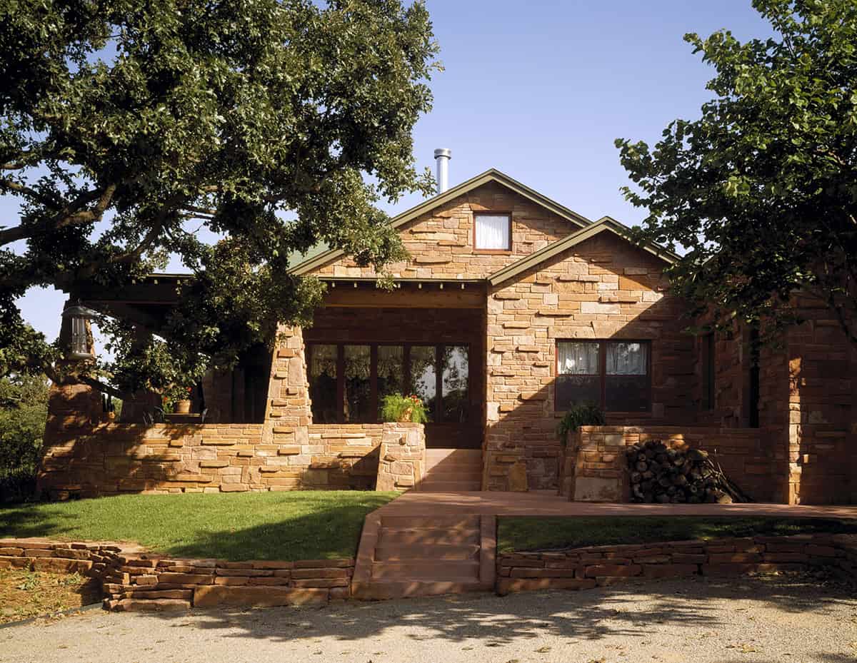 Front view of REIF Residence, a rustic Oklahoma home with native stone walls, green diamond shingles, and elevated porch by Rand Elliott Architects.