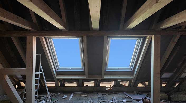 Zwei Dachfenster in einem hölzernen Dachstuhl mit Blick auf den blauen Himmel.