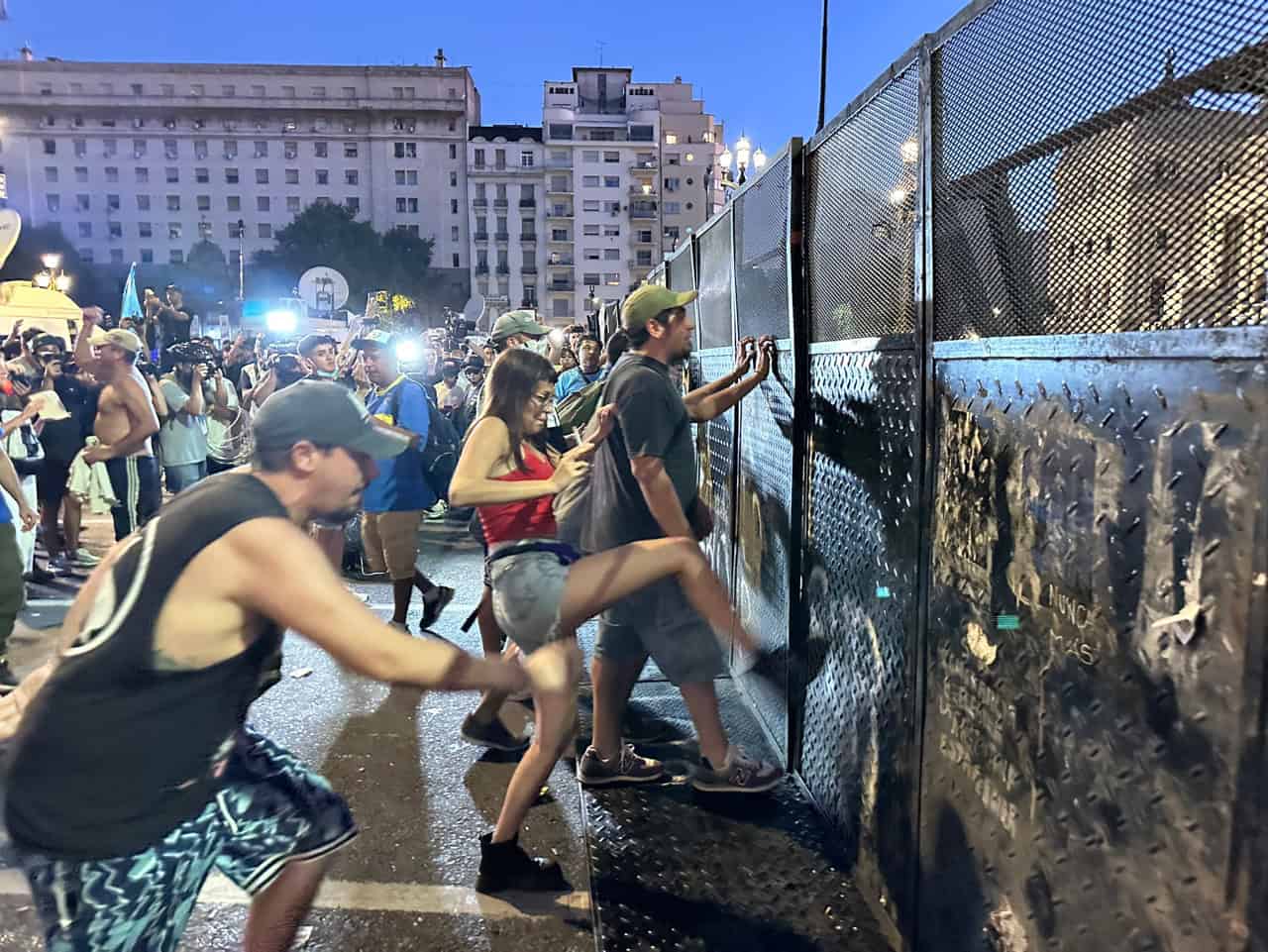 Protesters push and kick barricades outside Argentina’s National Congress as night falls, illuminated by streetlights and camera flashes.