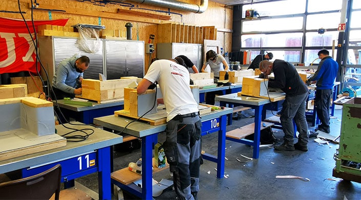 Bild von Azubis in der Werkstatt des Bildungszentrums Mehrere Personen arbeiten in einer Werkstatt an Werkbänken, tragen Arbeitskleidung und konzentrieren sich auf handwerkliche Tätigkeiten.